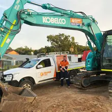 Man in safety gear standing beside a Kobelco excavator at a construction site with temporary barriers in the background.